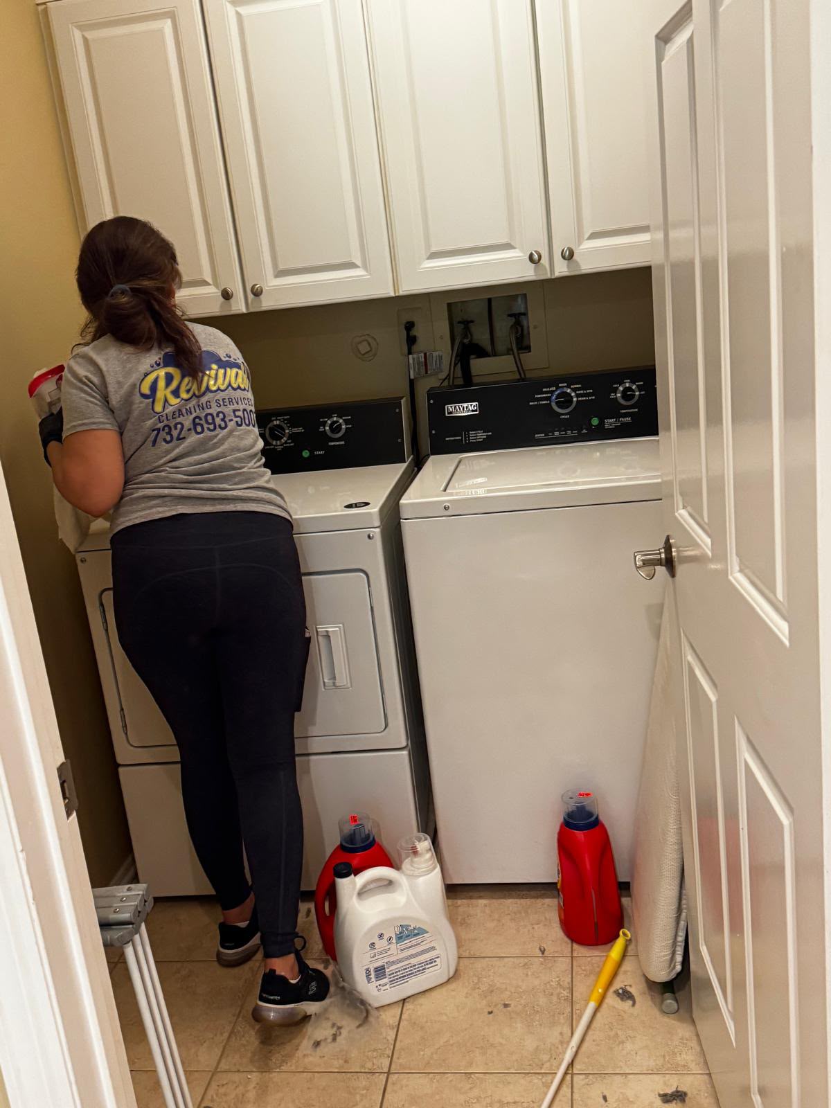 A Revival Cleaning Services team member in a branded uniform wiping down the washer and dryer in a home laundry room
